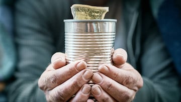 The worn hands of a homeless man hold out a collection can for a donation.