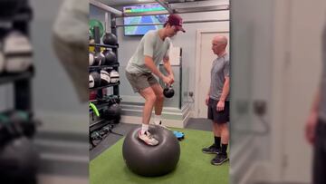 El snowboarder Scotty James con una kettlebell y una fitball entrenando el equilibrio en un gimnasio, con su entrenador mirando, material y una televisión al fondo.
