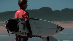 Un surfista vestido con la licra del Campeonato de Espa&ntilde;a de Surf 2020, mirando al agua con su tabla de surf bajo el brazo. 