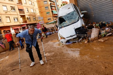 Un hombre con muletas camina junto a un coche averiado, tras las fuertes lluvias en Alfafar.