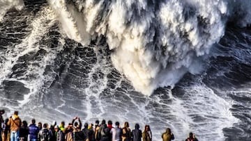 Una montaña de agua de una ola gigante rompe frente a varios aficionados que miran una sesión de surf XL desde un acantilado en Praia do Norte, Nazaré (Portugal).