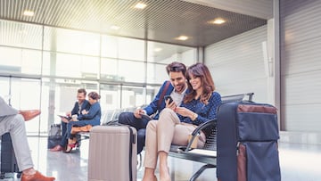Young businessman and businesswoman sitting on bench at airport lounge using mobile phone, with people in background. People waiting for flight. PAREJAS AEROPUERTO