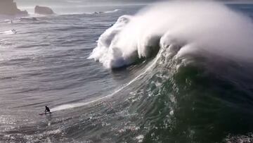 Una ola gigante rompiendo sobre un surfista en Praia do Norte (Nazaré, Portugal), con un jet ski y los acantilados al fondo.