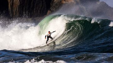 Un surfista surfea una ola que rompe detrás suyo y que mide dos metros en Tapia de Casariego (Asturias, España), con las montañas al fondo.