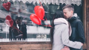 Una pareja besándose frente a un escaparate mientras sujetan unos globos con forma de corazón