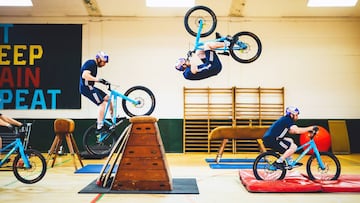 Danny MacAskill realiza un truco con su bici de MTB aprovechando un trampolín, un plinton y una colchoneta de un gimnasio.