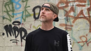 El skater Keith Hufnagel con una sudadera negra de HUF Worldide con un piano dibujado en el brazo y el logo en el pecho. Al fondo, una pared con varios graffitis.