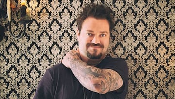 Bam Margera, de cintura para arriba y frente a una pared tapizada, toc&aacute;ndose el hombro y mirando a c&aacute;mara. 