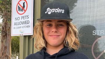 Khai Cowley, surfista, sonriendo, con gorra, frente a un cartel de que no se admiten mascotas.