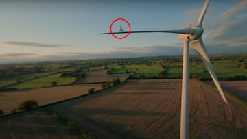 El piloto de MTB Danny MacAskill (dentro de un círculo rojo) montando en bici sobre la hélice de una torre eólica con campos de conreo debajo y un cielo seminublado al fondo.
