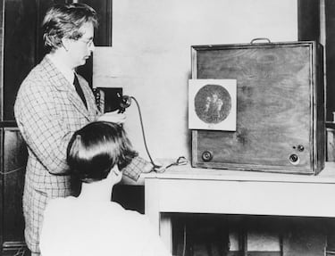 John Logie Baird realizando una demostración de su estación receptora, alrededor de 1927. «La pantalla redonda en la parte frontal del gabinete muestra a un asistente sosteniendo muñecos, mientras Baird dirige los movimientos en la pantalla por teléfono». y desarrolló un rudimentario aparato de televisión, capaz de transmitir y recibir imágenes en un radio de unos pocos metros. La primera demostración real se realizó en dos habitaciones abuhardilladas en el Soho de Londres, a principios de 1926, y para 1927 logró transmitir imágenes por línea telefónica desde Londres a Glasgow.
