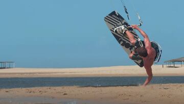 El kitesurfista Manel Arpa, sin camiseta y en ba&ntilde;ador, practicando kitesurf freestyle con una mano en la arena en un spot de Brasil. 