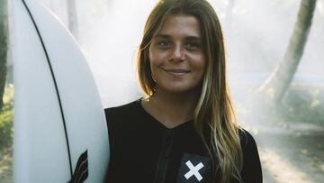 La surfista Daniela Boldini posa con un neopreno negro de Blue Banana y con su tabla de surf en un bosque de palmeras de las Islas Mentawai (Indonesia), con el sol entrando por los árboles. En verano del 2022.