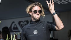 Matt Wilkinson, surfista australiano rubio con gafas de sol y traje de neopreno, levantando el brazo para saludar a alguien durante un evento de la Wordl Surf League en Australia.