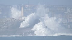 Olas gigantes rompiendo con fuerza contra un faro en Cantabria, Espa&ntilde;a, con la ciudad de Santander al fondo. 