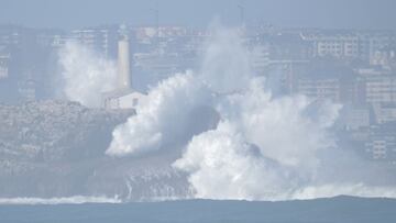 Olas gigantes rompiendo con fuerza contra un faro en Cantabria, España, con la ciudad de Santander al fondo.