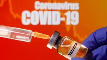 A small bottle labeled with a &quot;Vaccine&quot; sticker is held near a medical syringe in front of displayed &quot;Coronavirus COVID-19&quot;.