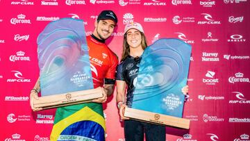 Gabriel Medina y Caroline Marks sujetan sus trofeos de campeones del Narrabeen Classic presented by Corona del Championship Tour (CT) de la World Surf League (WSL), con un fotocall rojo de fondo. En abril del 2021.