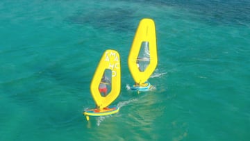 Dos windsurfistas navegan con las nuevas tablas y velas 100% hinchables Tamahoo 100 de Decathlon, de color amarillo, en aguas cristalinas de un mar calmado y con una peque&ntilde;a isla al fondo a la izquierda. 