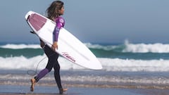 La surfista Marcela Machado de camino al agua con su licra morada y su tabla de surf en la SuperLiga Siroko. Con olas de fondo.