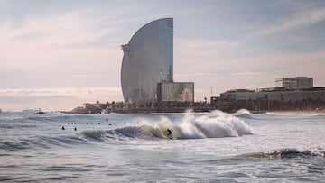 Un surfista surfeando una ola en la playa de la Barceloneta, con el hotel W al fondo y un cielo despejado, con nubes altas. 
