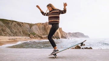 Chica patinando en longboard frente a una playa y unos acantilados. 