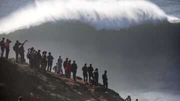 Una ola gigante rompiendo en Praia do Norte (Nazaré, Portugal) con varioespectacores mirándoselo desde el acantilado.