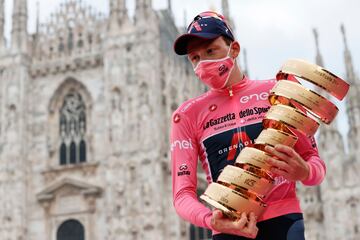 Tao Geoghegan Hart celebrando en el podio su victoria en el Giro de Italia, con la camiseta rosa de líder, sostiene el trofeo frente a la catedral del Duomo,