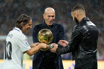 Modric y Zidane hacen entrega del Balón de Oro a Benzema, en un breve acto bajo la lluvia, el pasado sábado.