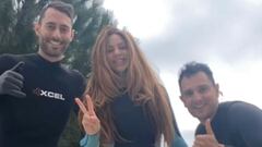 Shakira haciendo el s&iacute;mbolo de la victoria junto a dos surfistas haciendo el shaka t&iacute;pico del surf, todos con neopreno, con el cielo nublado de fondo y una playa de Sitges en la parte baja. 