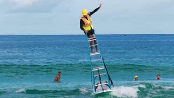 El surfista Jamie O'Brien, vestido de obrero con peto y casco, sobre una escalera en una tabla de surf surfeando una ola en Hawái.