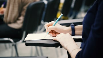 A girl writes a dictation or fills out documents in the audience, sitting on a school chair with a writing stand. Close-up. No face