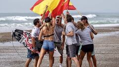 Gony Zubizarreta, Leti Canales, Andy Criere y otros miembros de la selecci&oacute;n espa&ntilde;ola de surf chocando las manos con dos banderas espa&ntilde;olaen una playa, con olas al fondo. 