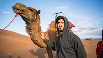 El surfista y emprendedor Tono García, con una sudadera gris y la capucha puesta, acaricia un camello en el desierto de Marruecos.