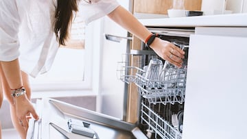 Woman with long hair in white blouse puts crockery to wash in the dishwasher at home.