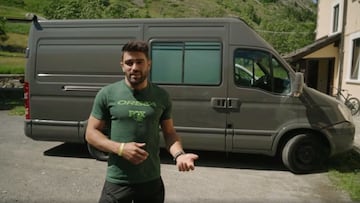 El rider Edgar Carballo habla con una camiseta verde de Orbea frente a su furgoneta de viaje, con una bici de MTB al fondo, apoyada en su casa; y montañas con árboles en la otra parte del fondo.