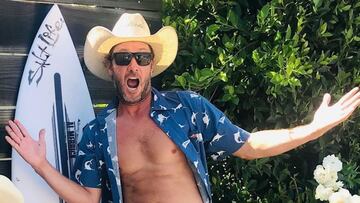 Tim Curran, surfista, abriendo los brazos, con gafas de sol y sombrero de vaquero, con su tabla de surf al fondo.