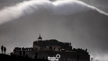 La ola gigante de Nazaré conocida como big mama rompiendo justo frente al faro y el fuerte de San Miguel (Nazaré, Portugal) ante una multitud congregada para observar el espectáculo.