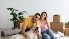 Full length portrait of happy young diverse couple with their dog posing on floor of new home on moving day. Millennial homeowners with cute pet sitting among cardboard boxes, relocating to new house