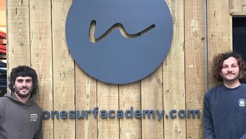Los surfistas Toni Varela y Carlos Fernández apoyados en una pared de madera con el logo y las letras de One Surf Academy.