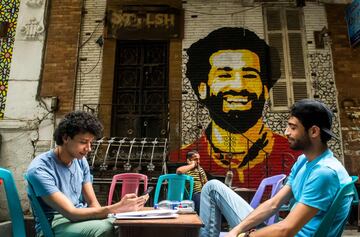 Jóvenes sentados en una terraza de una cafetería de El Cairo en la que hay un mural pintado con el rostro de Mohamed Salah. 
