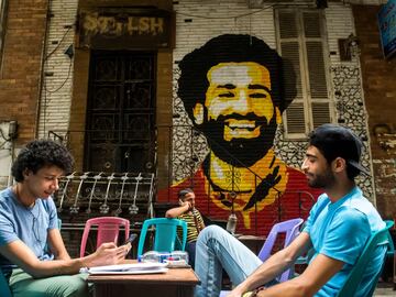 Jóvenes sentados en una terraza de una cafetería de El Cairo en la que hay un mural pintado con el rostro de Mohamed Salah. 
