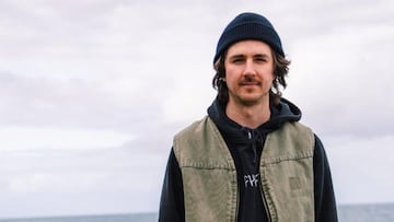 El rider de BMX Corey Walsh, con bigote y el pelo saliéndole por debajo del gorro, medio sonríe a cámara vestido con una sudadera de Cult, con el mar de fondo y el cielo nublado.