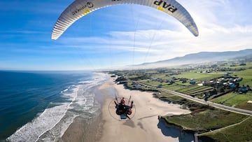 Nico Aubert sobrevolando una playa de España con olas y casas con césped al otro lado. En paramotor. En noviembre del 2023.