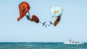 Dos riders de Wingfoil saltan de manera coordinada en la playa. 