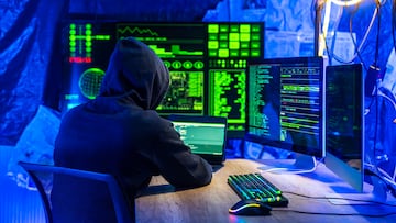 Un hacker encapuchado está programando en un portátil y varias pantallas de ordenador, intentando vulnerar la seguridad de la red y robar información sensible en una imagen teatralizada.