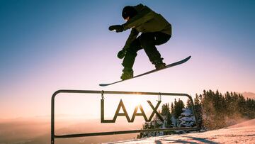 Un snowboarder salta sobre un r&oacute;tulo de la estaci&oacute;n de esqu&iacute; Laax, en los Alpes (Suiza), con unos abetos nevados y una puesta de sol de fondo. 