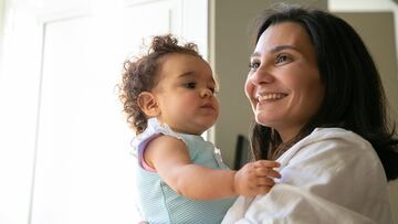 Cheerful mom holding sweet baby daughter in arms. Cute curly haired little girl looking at mother. Parenthood and childhood concept