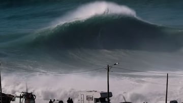 Una ola gigante a punto de romper en Praia do Norte (Nazaré, Portugal), vista desde el acantilado, con varios coches y autocaravanas, así como los palos de luz que hay.