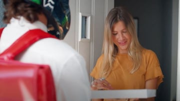 El rider de MTB Theo Erlangsen, con casco y en primer plano desenfocado, entrega una pizza a domicilio a una chica rubia que está a punto de abrirla en la puerta de casa.